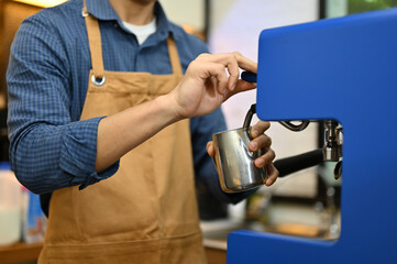 Cropped image of a barista in apron steaming fresh milk with espresso machine, working in cafe.