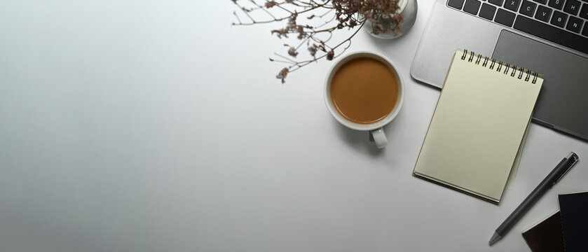 Top view of empty notepad, cup of coffee, laptop and dried flowers on white table. Copy space for your text