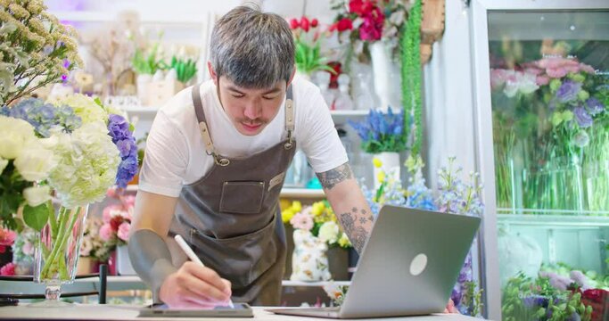 Asian male florist shop owner in apron looking at laptop making order and writing down in tablet. Tattooed guy hipster seller in floral store working on computer. SME Small business man entrepreneur.

