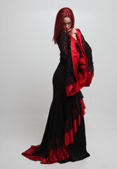 Naklejka premium portrait of beautiful red haired woman wearing long black fantasy vampire costume gown, isolated pose on studio background.