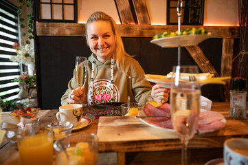 Smiling woman holding a glass of prosecco while sitting at a decorated breakfast table with a selection of cold cuts and healthy salads in foreground