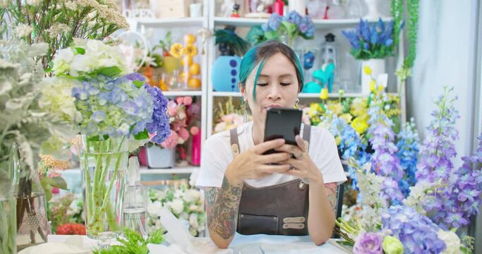 Asian female florist shop owner in apron working in store. Tattooed woman hipster seller taking picture for customer and sending photo to client. E-commerce, SME entrepreneur, Small business online.
