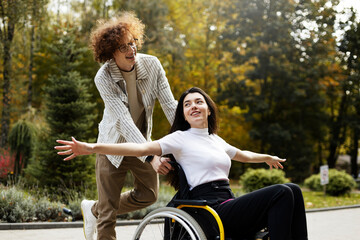 Cheerful, smiling guy is driving a wheelchair with a girl outdoors. A cheerful girl spread her arms to the sides, like the wings of an airplane, on the street. - Powered by Adobe
