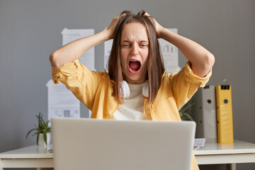 Portrait of shocked stressed depressed woman with brown hair working on laptop, having big problems, do not know what to do, screaming loud and pulling her hair, office background,