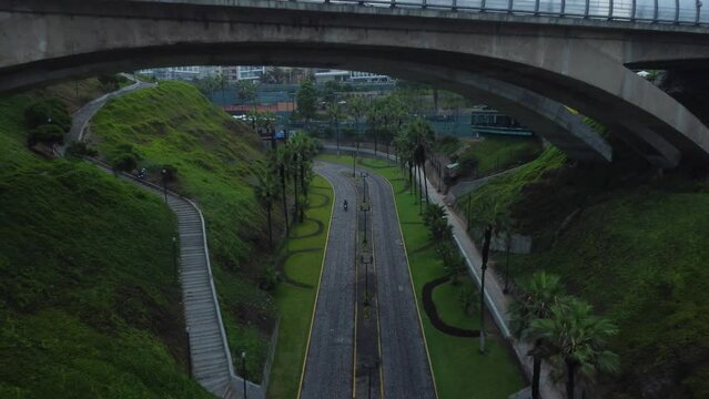 Street called "Bajada Balta" that goes under a bridge. Drone flies backwards showing the cityscape of many tall apartment buildings. Recorded early in the morning in Miraflores district of Lima, Peru.