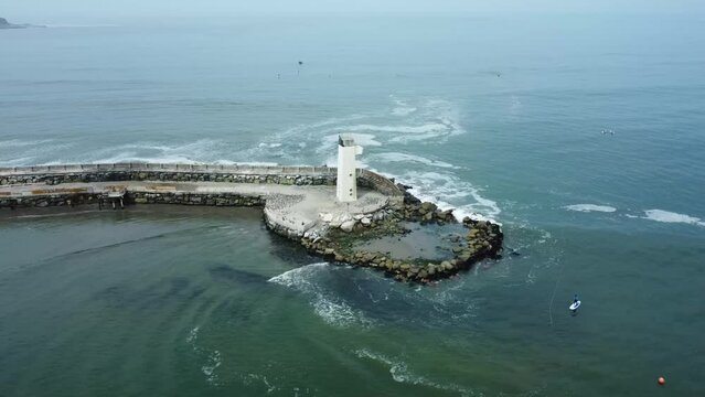 Drone orbits around a white small lighthouse in the edge of a rocky pier. In the background coastal mountain begin to appear as the camera turns left. Located in Lima, Peru.