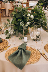 Wedding. Banquet. On the festive table with a tablecloth is a composition of flowers and greenery, there are plates with green napkins on wicker stands, glasses, cutlery and candles in glass vases