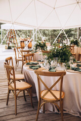 Wedding in the woods. Decor. The festive tables in the tent are decorated with compositions of flowers. On the tables plates, candles, glasses, napkins with cutlery