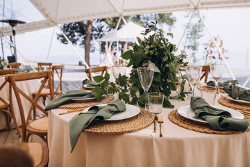 Wedding. Banquet. On the festive table with a tablecloth is a composition of flowers and greenery, there are plates with green napkins on wicker stands, glasses, cutlery and candles in glass vases