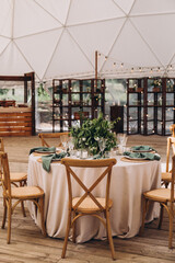 Wedding in the woods. Decor. The festive tables in the tent are decorated with compositions of flowers. On the tables plates, candles, glasses, napkins with cutlery