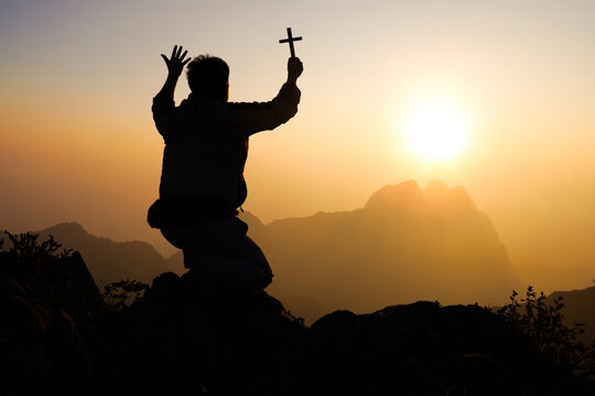  Silhouette of Young man  praying and holding christian cross for worshipping God at sunset background. Christian Religion concept background.