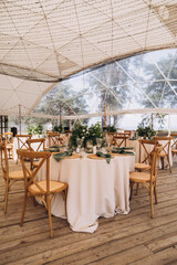 Wedding in the woods. Decor. The festive tables in the tent are decorated with compositions of flowers. On the tables plates, candles, glasses, napkins with cutlery