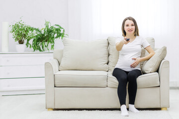 Obraz premium Cute pregnant woman sitting on sofa with remote control.