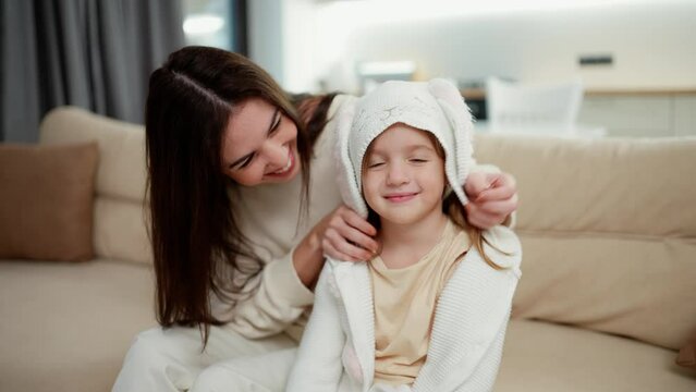 Loving mother embracing kid daughter and putting hood on the head while sitting on sofa