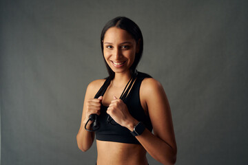 Fototapeta na wymiar Portrait of cheerful young biracial woman in sports bra holding jump rope over shoulders in studio