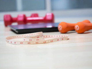 Closeup of measuring tape with blurry orange, red dumbbells,  weight scale on wooden floor
