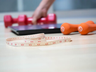 Closeup of measuring tape with blurry orange, red dumbbells,  weight scale on wooden floor and women 's hand for dieting or healthy concept