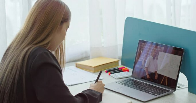 Young girl college student watching, studying lesson online from home. Woman taking notes on tablet while looking at computer following professor doing math study for class assignment. Rear back view.