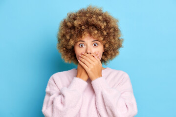Fototapeta na wymiar Shocked speechless woman with curly hair covers mouth tries to be silent received bad news looks shocked and amazed wears pink pullover isolated over blue wall