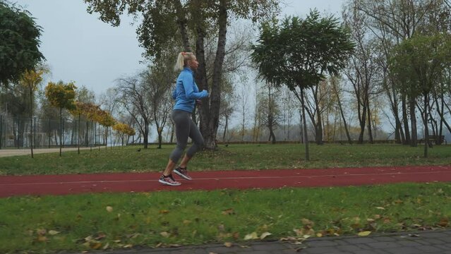 Young blonde woman wearing blue sports jacket jogging at track in autumn morning. Adult female jogger running outside. Fallen leaves lying on ground. Concept of healthy lifestyle