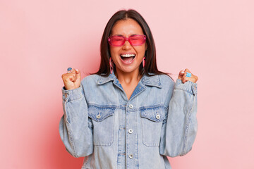 Charming emotional brunette caucasian woman clenches fists screams loudly celebrates own triumph wears transparent glasses and jeans jacket poses over pink background