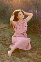 A cheerful young girl in a pink dress sits resting among lavender bushes and plays with her long hair. Sunset.