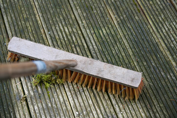 Outdoor broom with plastic bristles scrubbing a weathered wooden deck to remove algae and moss,...