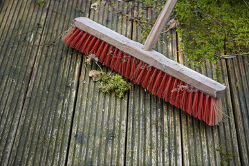 Red outdoor broom with stiff plastic bristles on a dirty wooden deck with algae and moss, spring cleaning in garden and yard for the new season, copy space, selected focus - 574211285