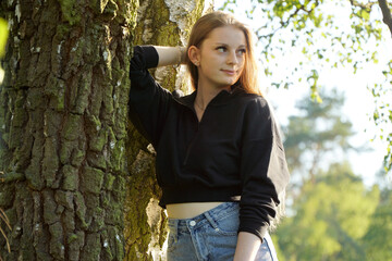 Beautiful young woman in hot pants and black jacket leans against a tree and smiles kindly on a sunny summer day in the park	
