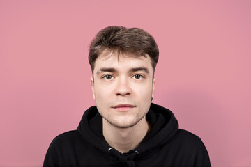 Obraz premium Portrait of a real handsome, pensive young brunette with brown eyes and an ear piercing on a pink background, looking directly at the camera