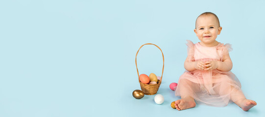 Happy baby girl, ten months old, wearing bunny ears headband, sitting on light blue studio background and playing with colored easter eggs. Little Child. Easter greeting card. Baby easter bunny.