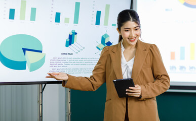 Millennial Asian happy professional successful female businesswoman presenter trainer standing holding touchscreen tablet presenting business ideas strategy graphs charts on monitor in meeting room