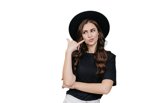 Brunette hipster girl in hat , black t-shirt makes call gesture by hand looks aside at copy space against transparent background. Caucasian fashion model shows sign call me. Mockup, promo, sale.