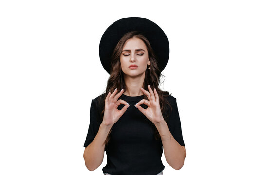 Confident brunette young woman in black hat with brim and black t-shirt eyes closed makes mudra shape in meditation stands over transparent backdrop. Relaxing hipster posing, thinks, makes choice