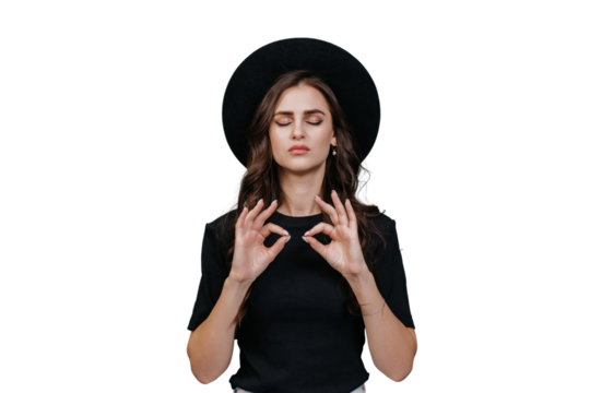 Confident brunette young woman in black hat with brim and black t-shirt eyes closed makes mudra shape in meditation stands over transparent backdrop. Relaxing hipster posing, thinks, makes choice - Powered by Adobe