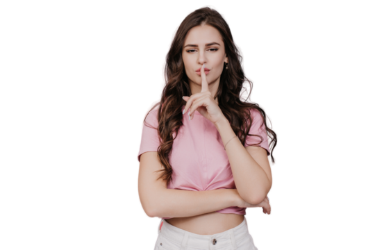 Confident young brunette with wavy hair touches lips bi index finger makes silence sign looks at camera stands against transparent backdrop. Pretty model in pink t-shirt has a beauty secret. Cosmetics
