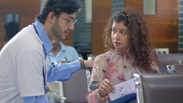 A helpful and supportive Indian Asian male doctor wearing an apron and stethoscope checking file, talking and consoling a sad and worried female relative of patient sitting in a hospital waiting area