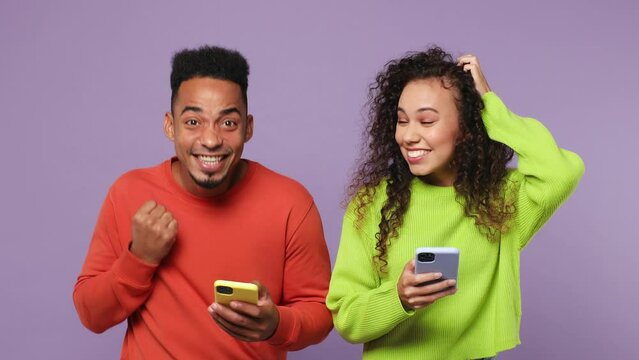Young couple two friends family man woman of African American ethnicity wear casual clothes together use mobile cell phone found out great big win news isolated on pastel plain light purple background