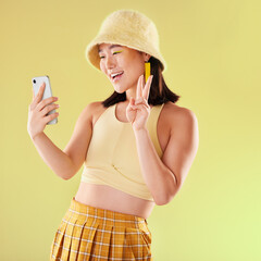 Selfie, peace sign and smile of Asian woman in studio isolated on a yellow background. Makeup,...