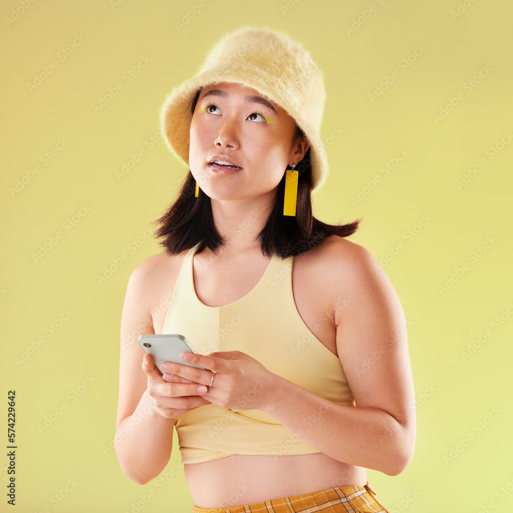 Wall mural Phone, thinking and Asian woman typing in studio isolated on a yellow background. Ideas, technology or female model contemplating, decision or lost in thoughts with mobile for social media or texting