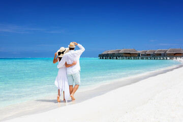 Happy couple in white clothing and with hats walks down a tropical beach with turquoise sea in the Maldives islands