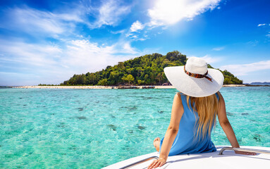 A beautiful tourist woman with sunhat sits on a yacht and looks at the turquoise sea and beaches of Bamboo island in the Krabi region, Thailand - obrazy, fototapety, plakaty