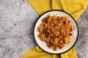 Homemade pilaf with rice, meat, carrots and onions on a white round plate on a dark gray background. Top view, flat lay