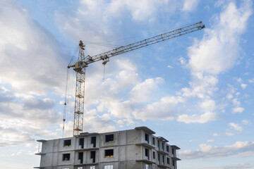 A large tower crane builds a house against a blue sky with clouds. Construction of an apartment building