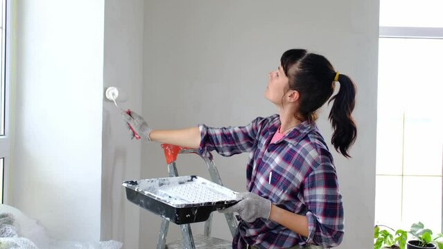 Woman in paint roller and white paint paints the wall of house on stepladder. Construction work and cosmetic repairs in house, wall painting, tinting, finishing work with your own hands