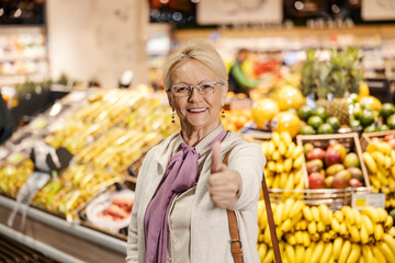 Portrait of a happy old woman is giving thumbs up while standing in hypermarket at fruit department and looking at the camera.