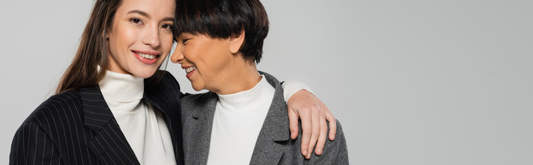 happy woman in black jacket hugging asian mother and smiling at camera isolated on grey, banner