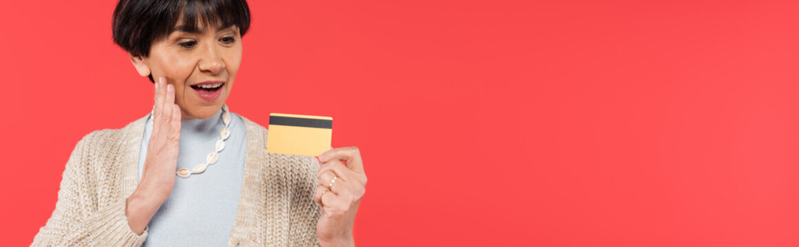 Impressed Asian Woman In Cardigan Touching Face While Holding Credit Card Isolated On Coral, Banner