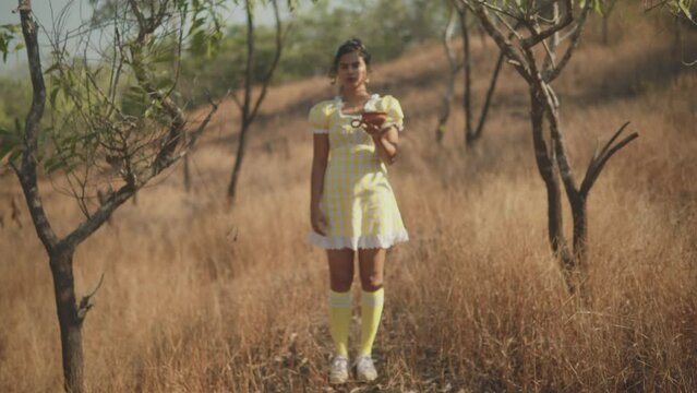 A shot racking focus on an Asian female wearing a pretty yellow dress holding a clay pot in the palm of her outstretched arm as she stands in a dry grass field on a summer&rsquo;s day, India 