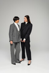 full length of cheerful asian mother and daughter in pantsuits looking at each other on grey